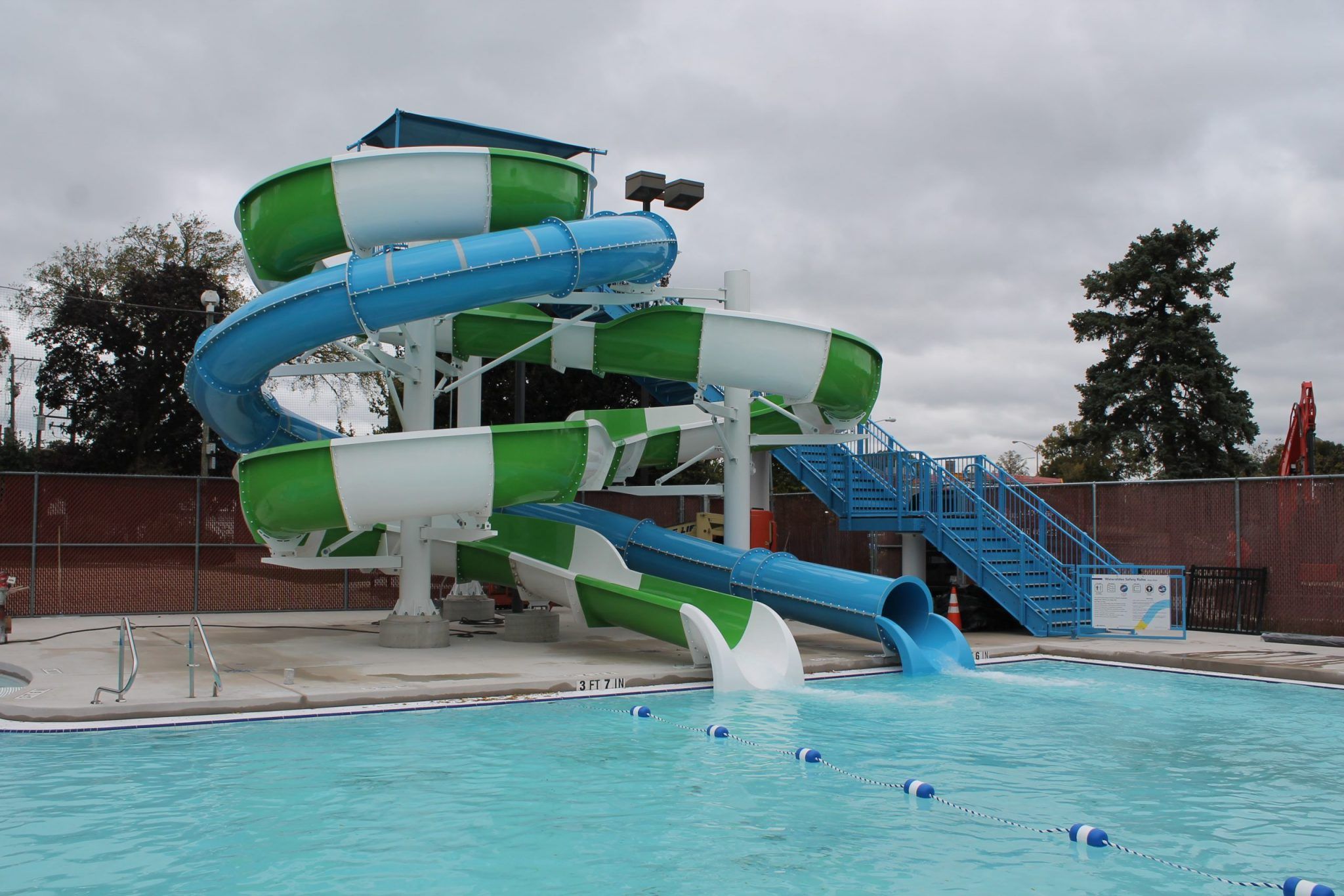A multi-level water slide complex with blue and green tubes, featuring a blue staircase leading to the top, emptying into a swimming pool at Rehm