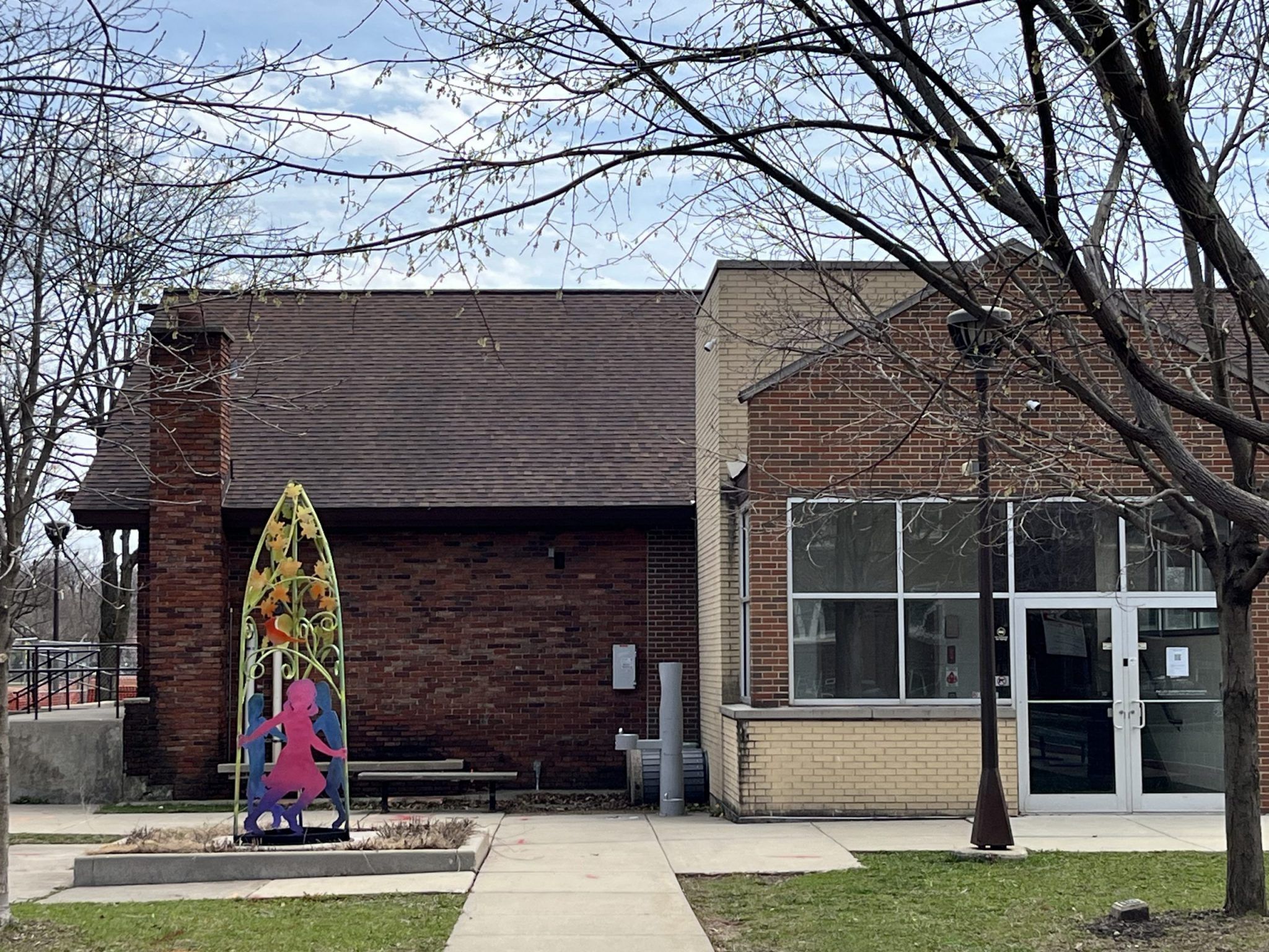 Colorful metal sculpture featuring silhouetted figures in front of a brick building at Longfellow Park.