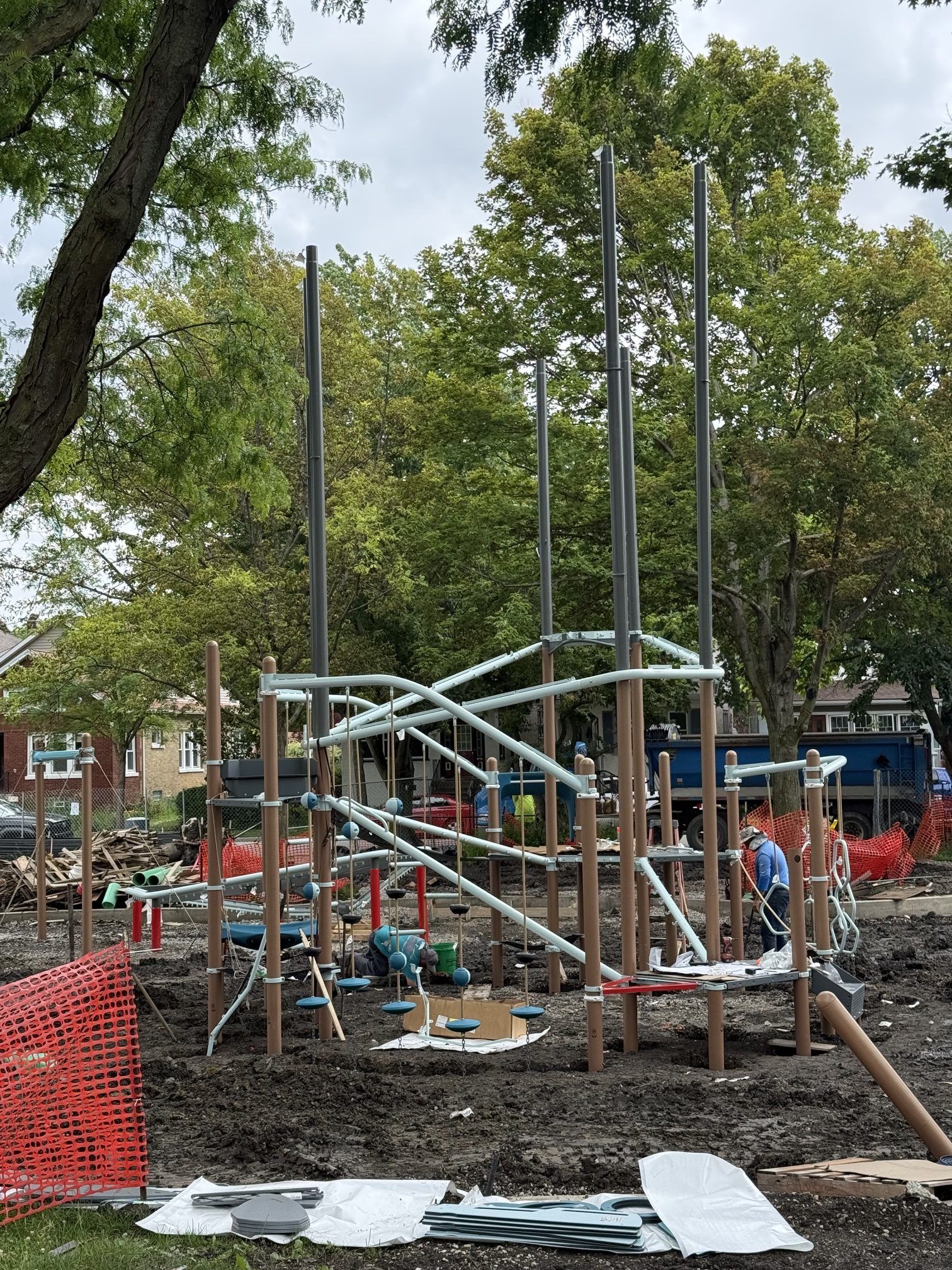 Construction of a new playground structure at Longfellow Park, featuring climbing elements and support poles. Workers are present, and safety netting