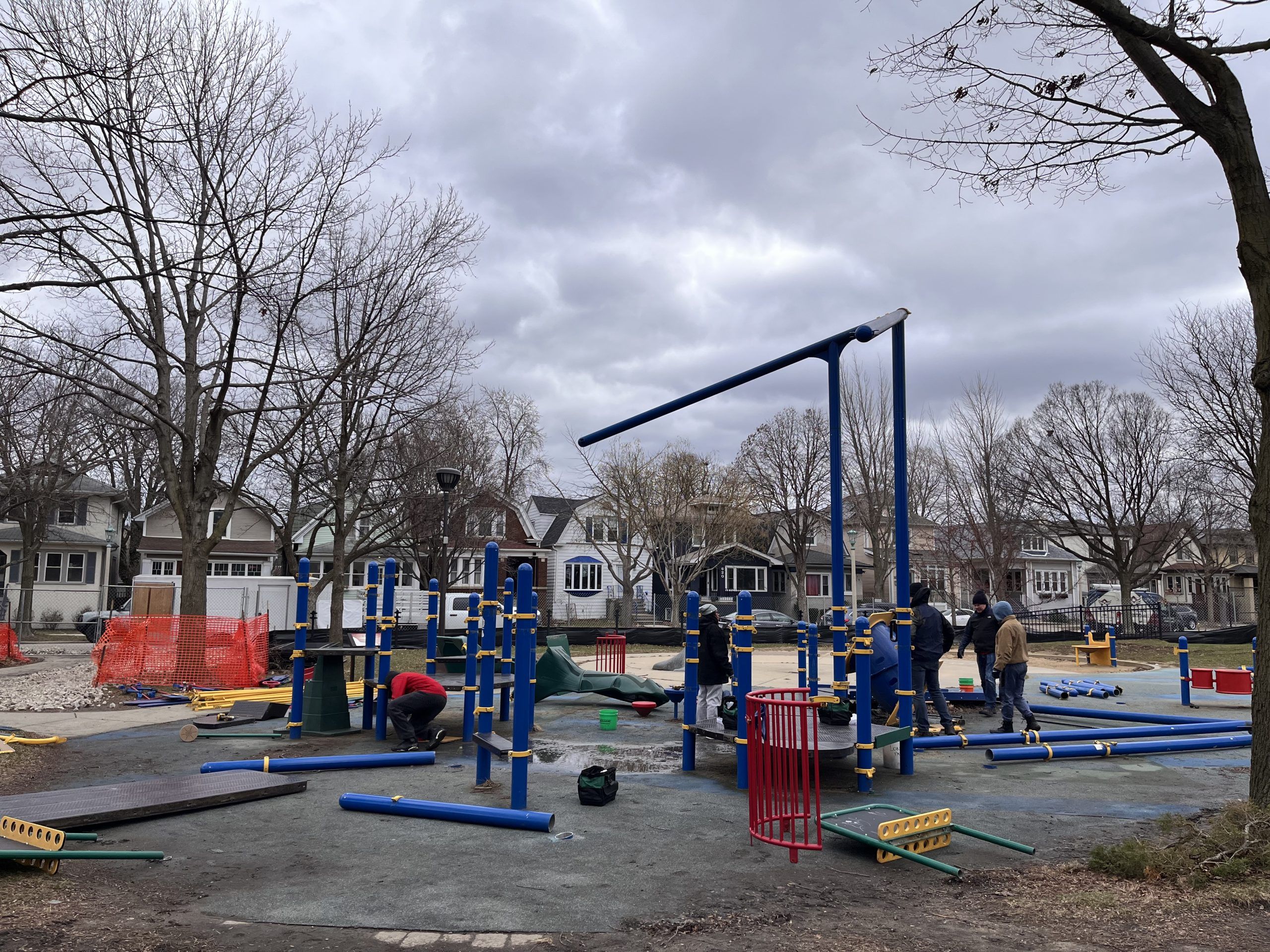 Construction workers assemble playground equipment at Longfellow Park under a cloudy sky. New blue and yellow structures are partially built.
