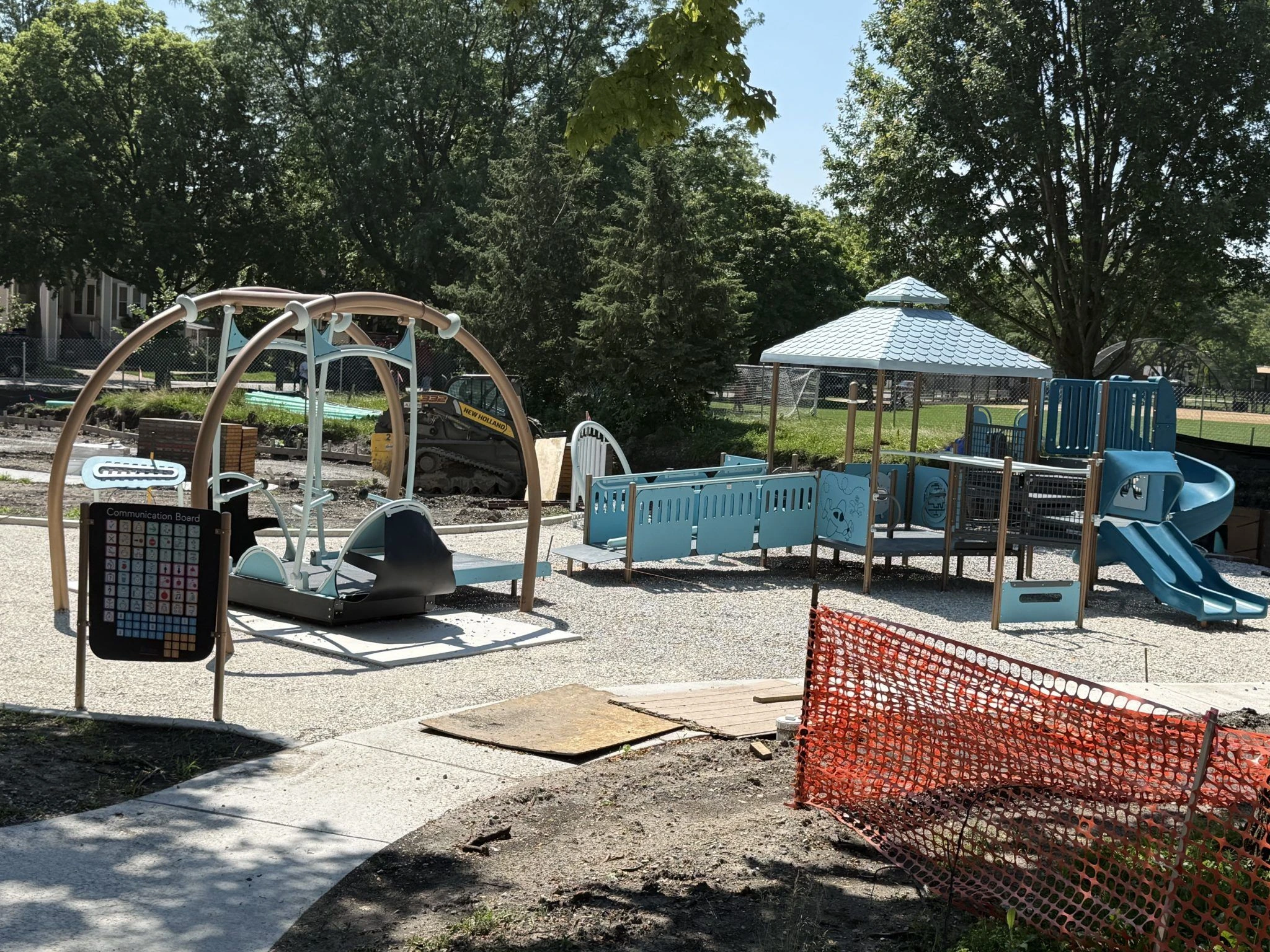 New playground equipment with blue accents and a communication board is installed at Longfellow Park. Construction fencing is visible.