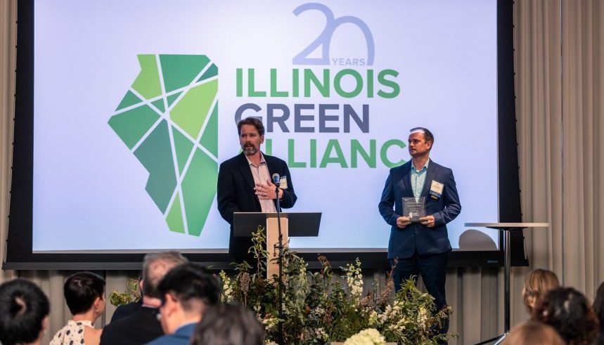 Two men stand on a stage in front of a screen displaying the "Illinois Green Alliance" logo and "20 Years." One man speaks at a podium, the other