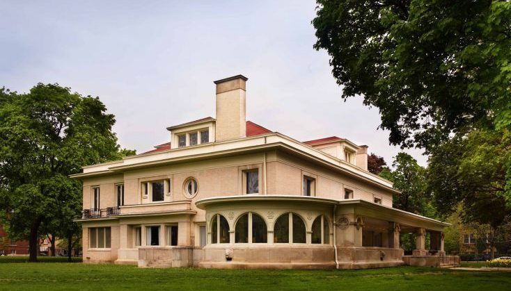A large, light-colored house with a red roof, featuring a rounded sunroom with arched windows and a prominent chimney. Surrounded by green lawn and