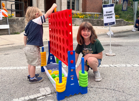 Two children play a giant Connect Four game outdoors. A girl smiles at the camera while a boy places a red disc.
