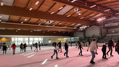 People ice skating on an indoor rink with a wooden ceiling and bright lights.