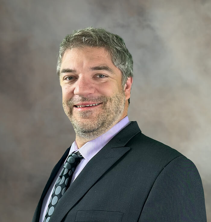 Jake Worley Hood, a man with graying hair and a beard, smiles wearing a suit and tie. He is part of the Board of Commissioners.