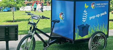 A three-wheeled cargo bike with a "pop-up park" trailer, featuring the Park District of Oak Park logo and graphics.