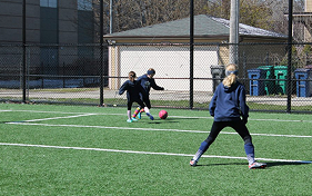 Three individuals playing soccer on an artificial turf field with white markings. One person is kicking the ball, while two others are positioned to