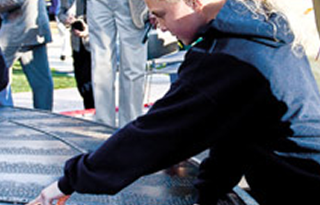 A person in a dark hoodie leans over a memorial plaque, touching the engraved names.