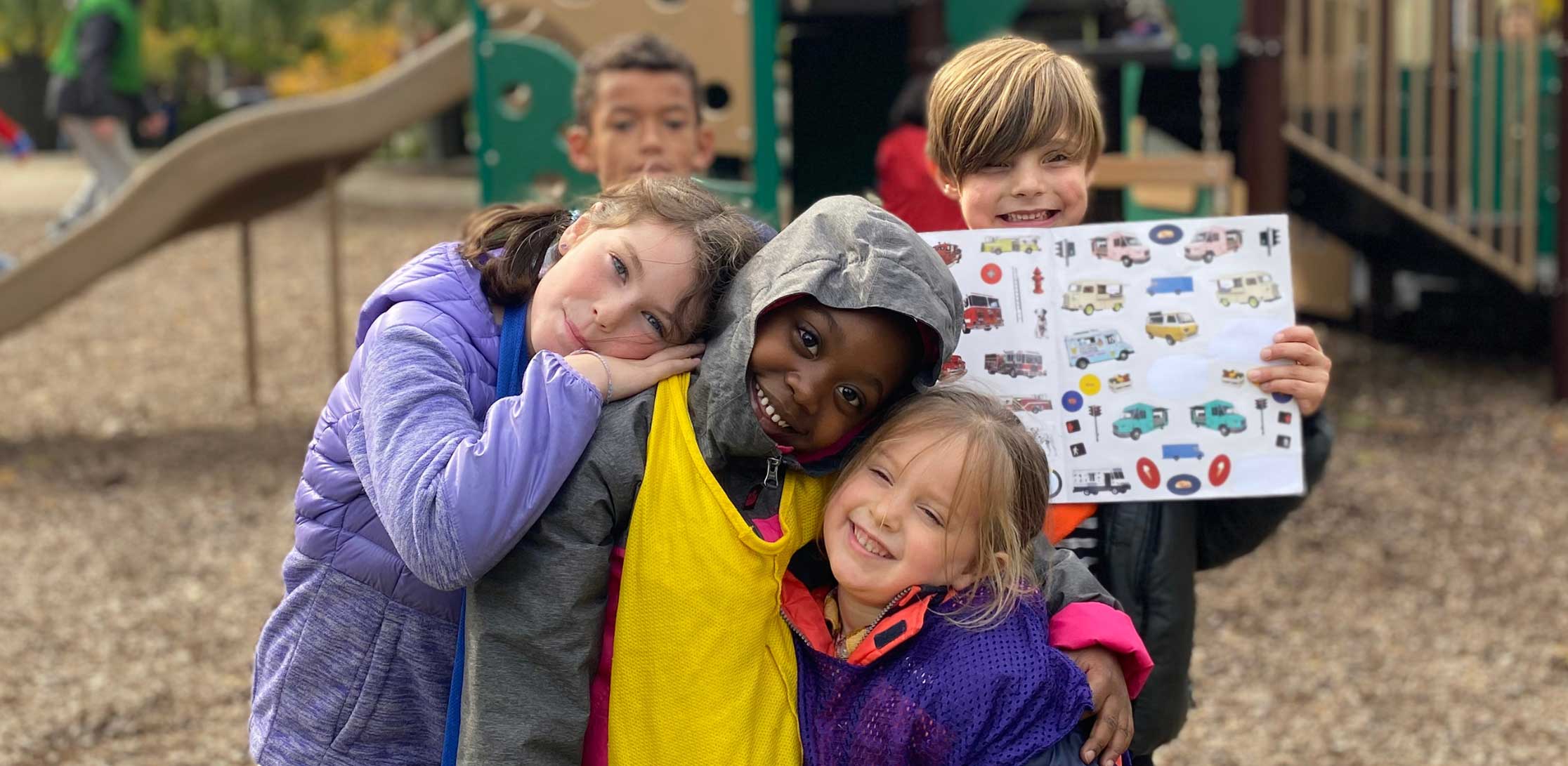 Four children smiling and hugging on a playground. One child holds up a sheet of vehicle stickers.