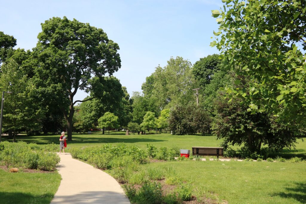 A paved path curves through a grassy park with mature trees. A woman in a red and white dress walks on the path.