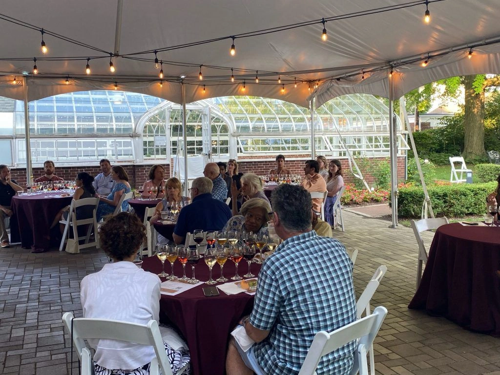 Guests seated at tables under a tent for a wine tasting event, with a greenhouse in the background.