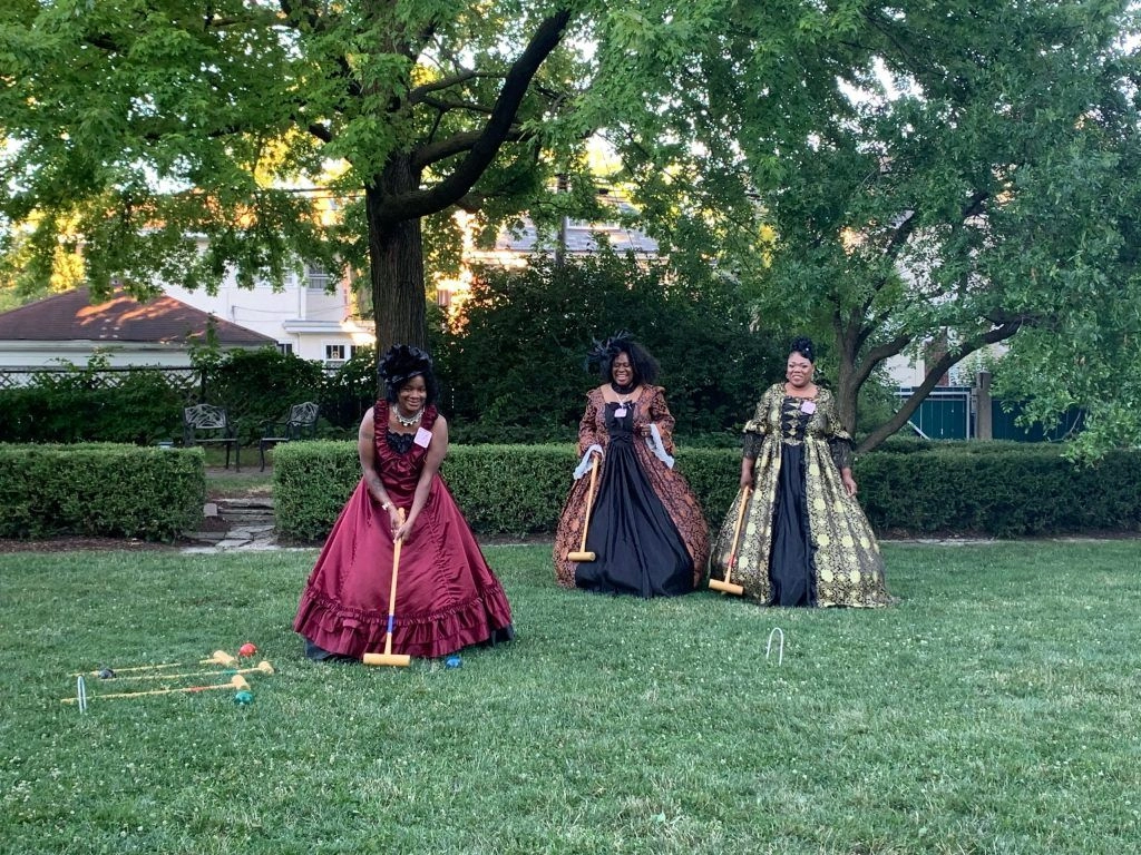Three women in historical gowns play croquet on a lawn.