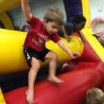 A young boy in a red shirt jumps on a red inflatable surface inside a bouncy castle, with other children visible.