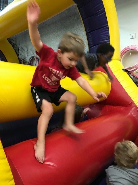 A young boy in a red shirt jumps on a red inflatable surface inside a bouncy castle, with other children visible.