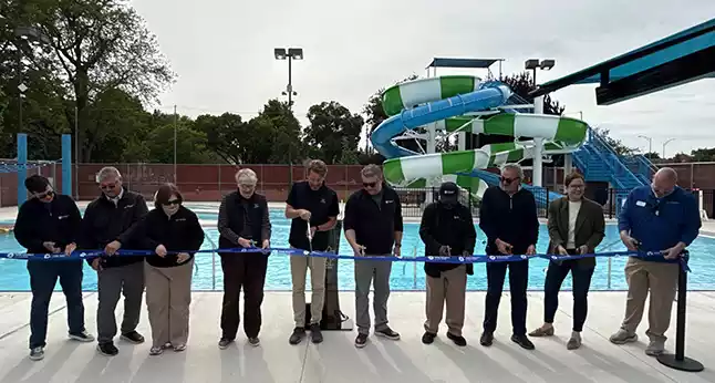 A group of people in business casual attire cut a blue ribbon in front of a swimming pool and water slide.