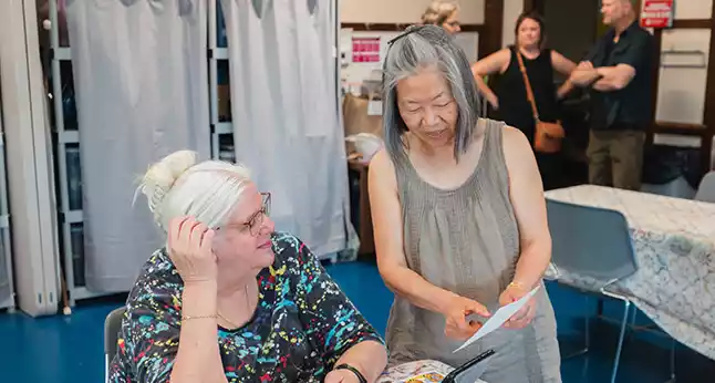 Two women, one with blonde hair in a bun and glasses, the other with gray hair, look at a document together.
