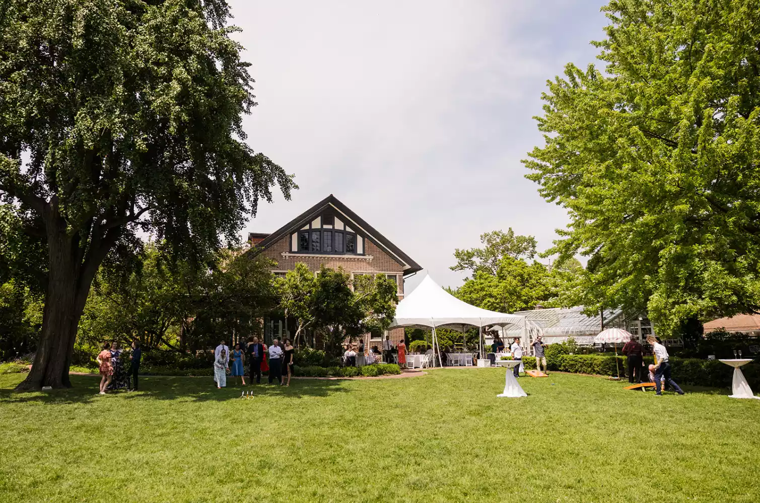 People gather on a lawn in front of a large house with a white tent set up for an event. Some guests mingle, while others play lawn games.