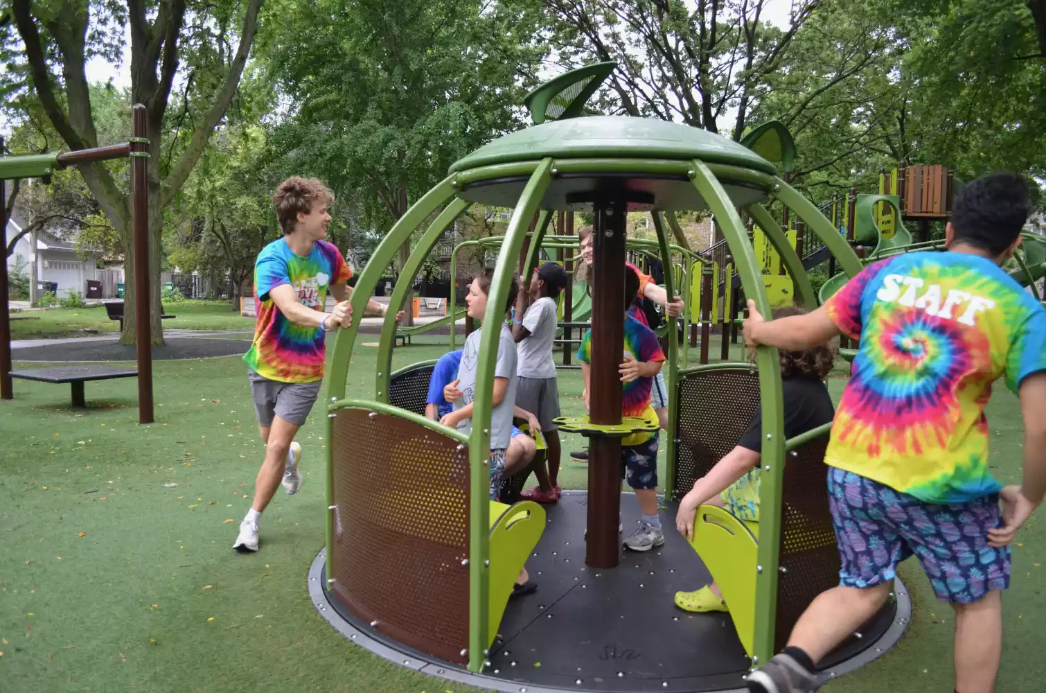 Several children and staff members wearing tie-dye shirts interact with a green merry-go-round at a playground.