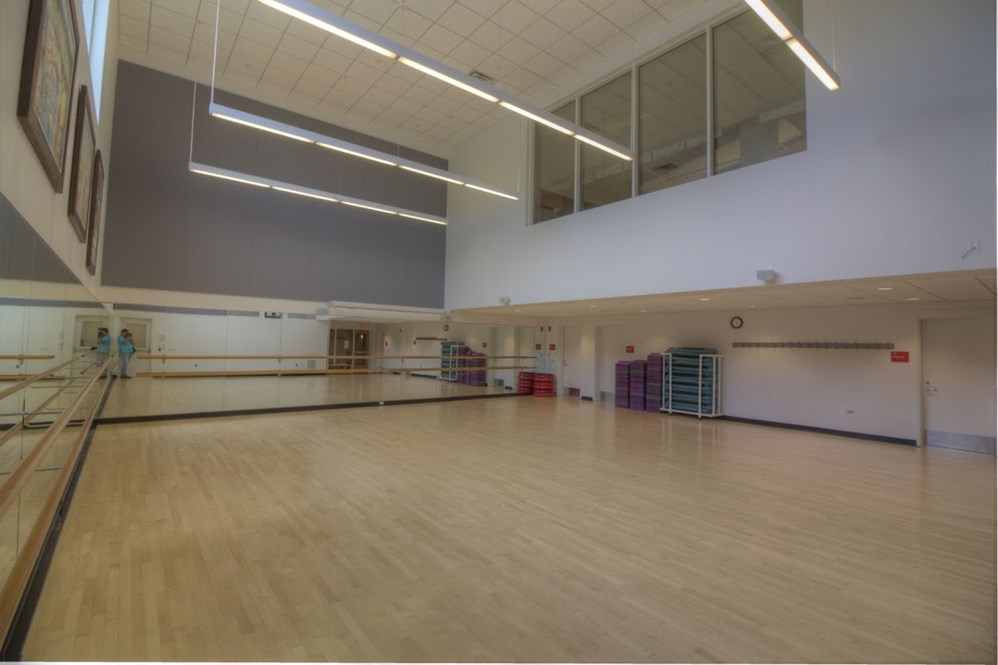 Spacious activity room with a mirrored wall, ballet barres, wooden floor, and stacked aerobic steps.