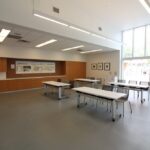 A spacious, well-lit medium activity room with multiple tables and chairs, a long counter with cabinets, and large windows.
