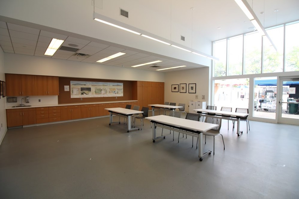 A spacious, well-lit medium activity room with multiple tables and chairs, a long counter with cabinets, and large windows.