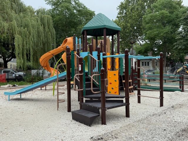 A modern playground structure with slides, climbing wall, and monkey bars, set on a gravel surface.