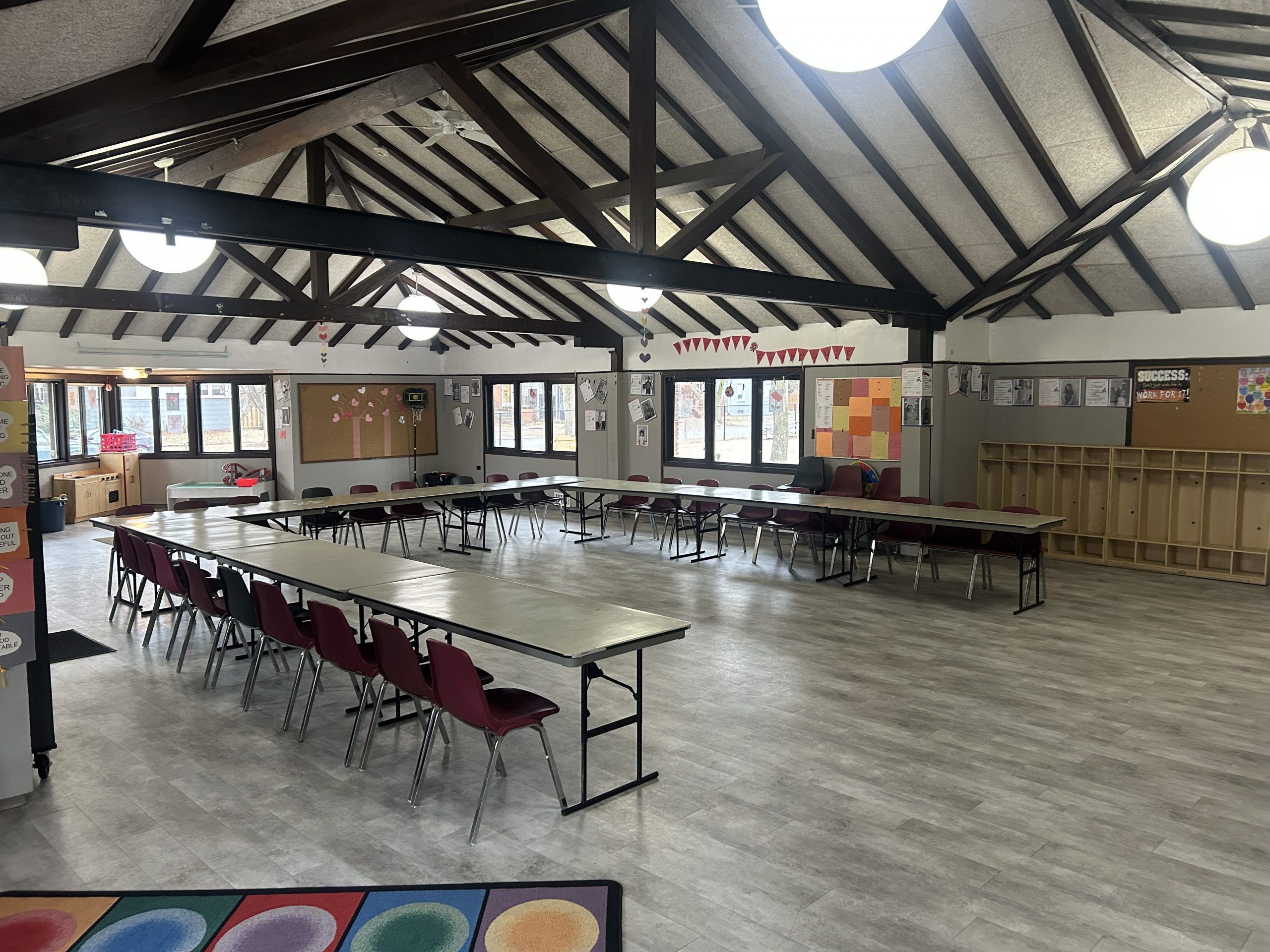 A spacious community room with U-shaped table arrangements, chairs, and wooden cubbies.