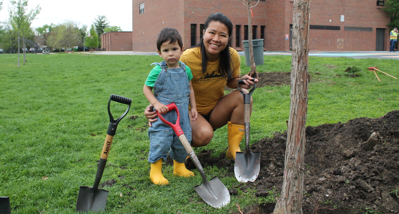 A woman and a young child kneel in the grass next to a newly planted tree, holding shovels. They are participating in Arbor Day tree planting.