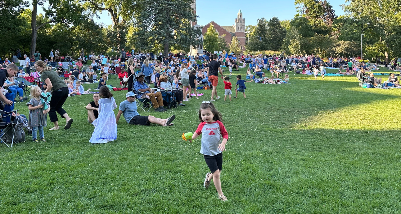 A young girl in a red and gray shirt runs across a grassy lawn towards the camera, holding a toy. Many people are gathered on blankets and in chairs