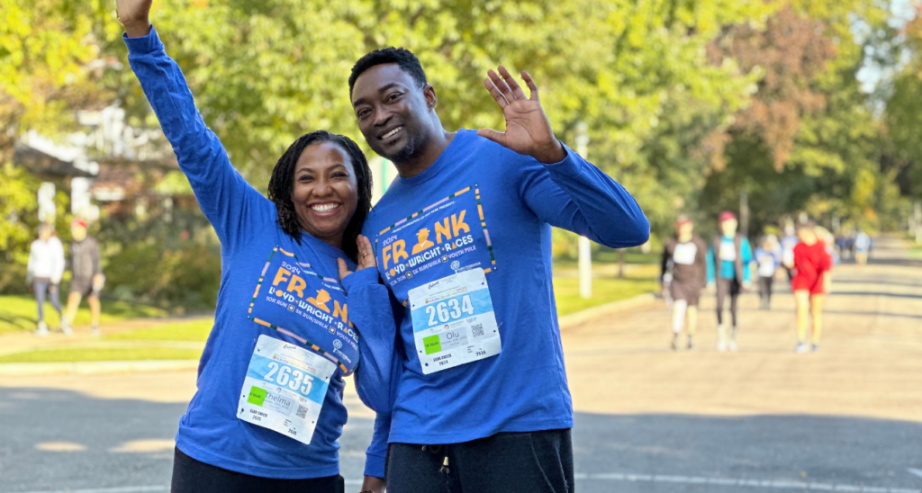 A smiling Black couple wearing blue long-sleeved shirts with "FRANK LLOYD WRIGHT RACES" and bib numbers 2635 and 2634. They are waving at the camera