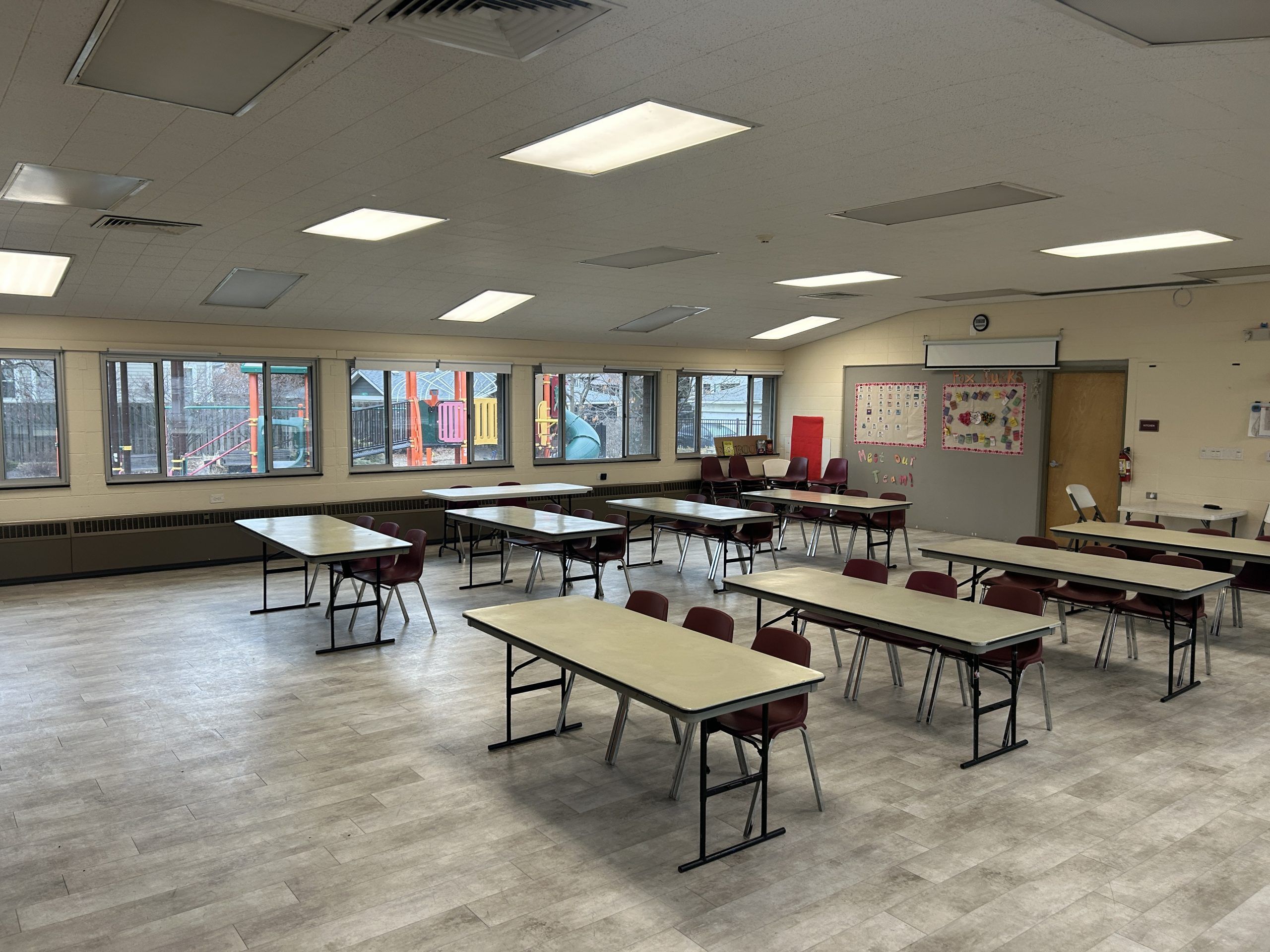 A community room set up with rows of tables and chairs, facing a wall with a projector screen and bulletin boards. Windows overlook a playground.