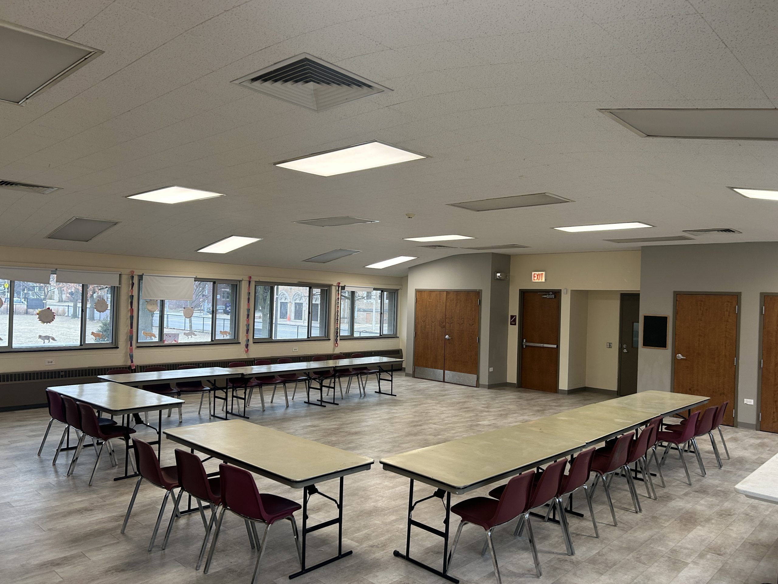 Rectangular tables and chairs are set up in a large room with windows overlooking an outdoor area. The room has an 'EXIT' sign above a doorway.