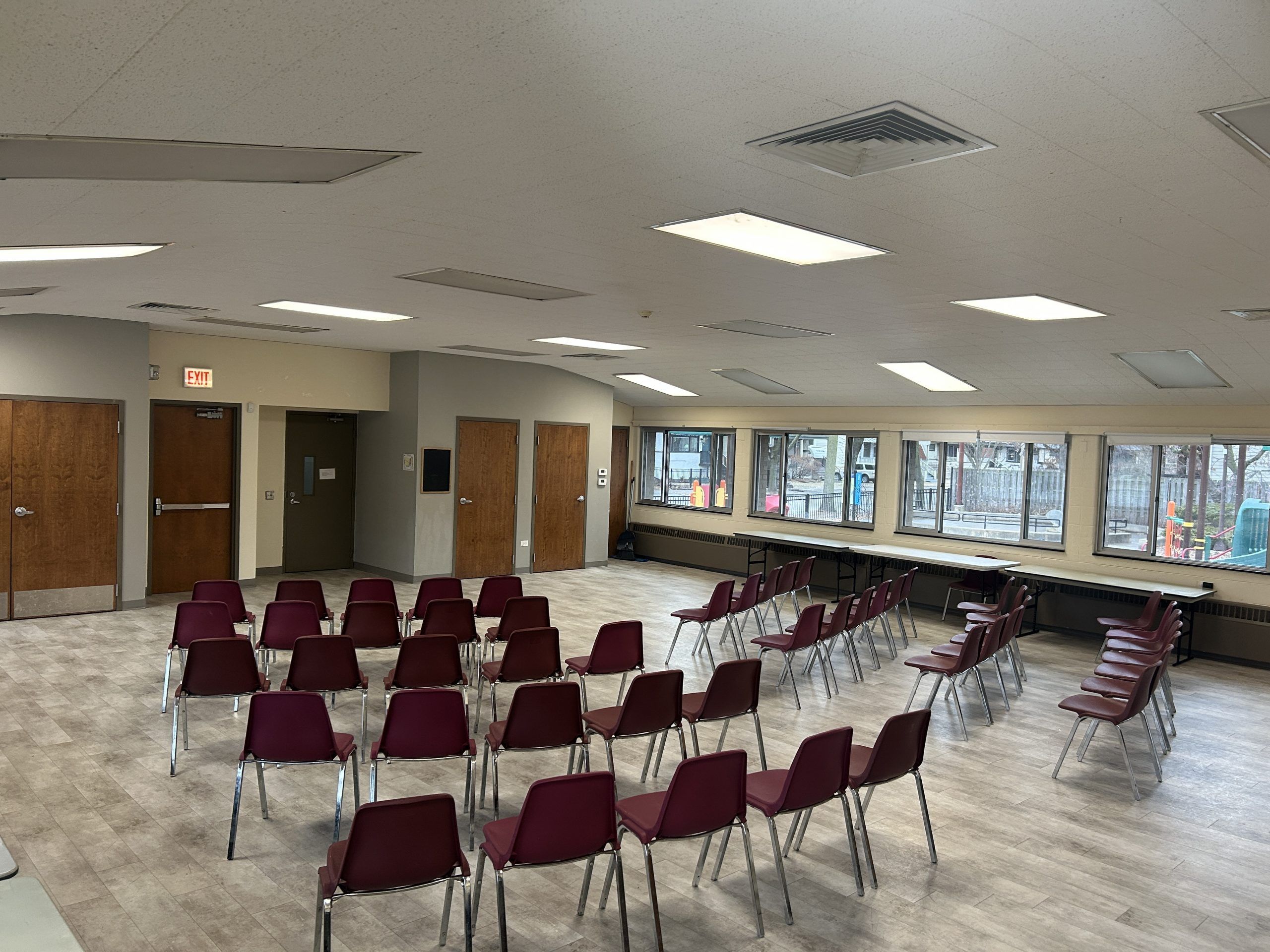 Rows of maroon chairs are set up in a large room with tables along the windows. An exit sign is visible above a door.
