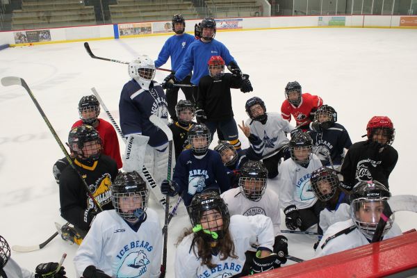 A group of young hockey players and coaches pose for a photo on an ice rink. Most players wear helmets and team jerseys.