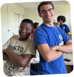 Two young men, one wearing a blue t-shirt and the other a tan t-shirt, stand with arms crossed and smile at the camera.