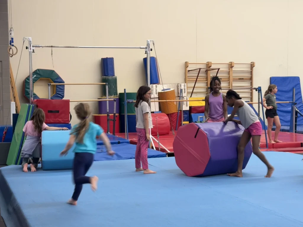 Young girls participate in gymnastics class, interacting with mats and equipment.