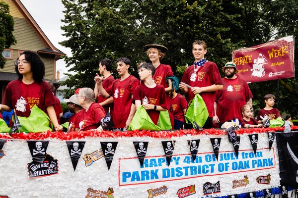 Group of people in red shirts on a parade float decorated with pirate flags and "Happy 4th of July from Park District of Oak Park" banner.