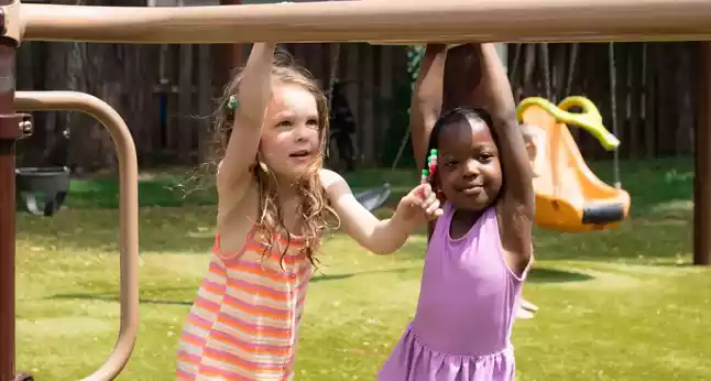 Two young girls, one with blonde curly hair and an orange and white striped dress, the other with dark skin and a purple dress, hang from a