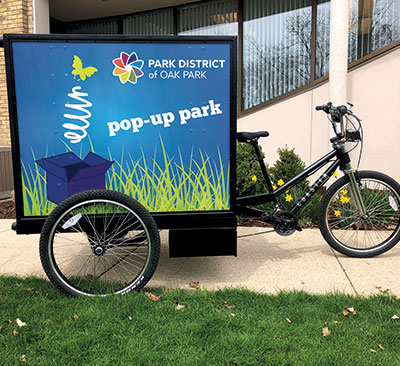 A three-wheeled bicycle with a large display box attached, advertising the "Park District of Oak Park pop-up park."