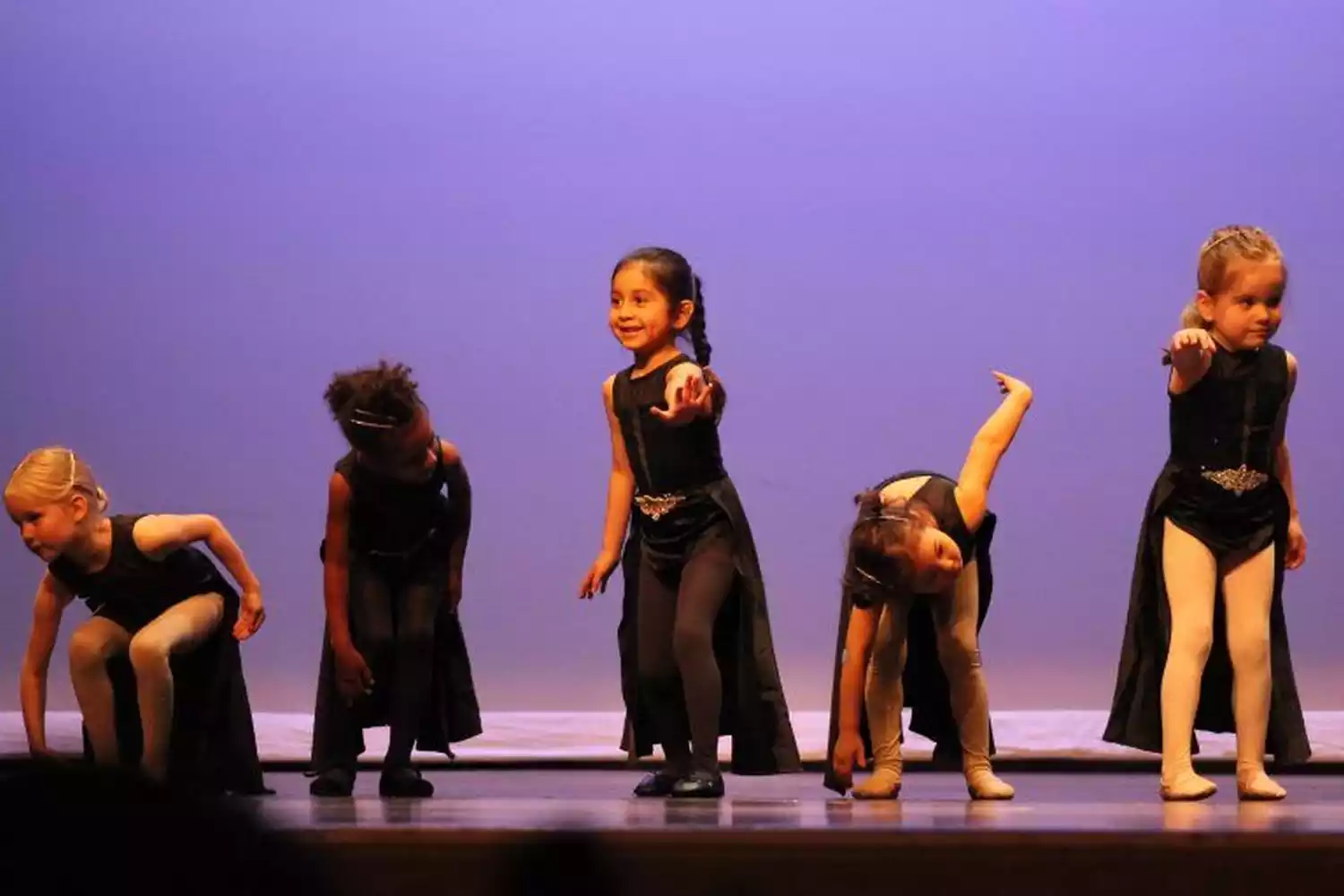 Five young girls in black dance costumes perform on a stage with a purple backdrop. They are in various poses, some bending and reaching.