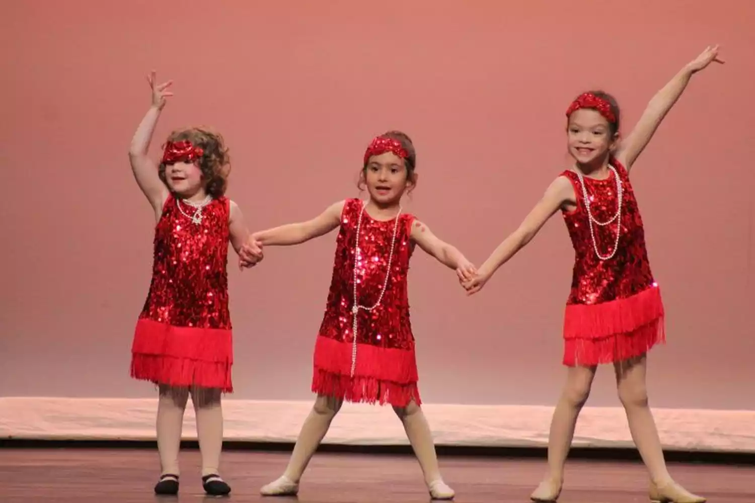 Three young girls in red sequined flapper dresses and headbands hold hands while dancing on stage.