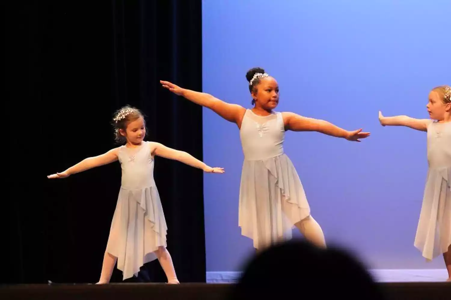 Three young girls in light blue ballet tutus perform a dance routine on stage, with arms outstretched.