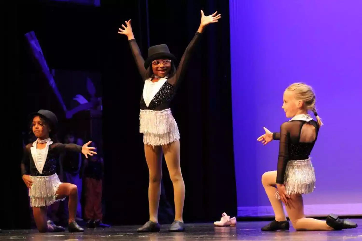 Three young dancers in sequined costumes and top hats perform on stage. The center dancer has arms raised, while the others are in kneeling poses.