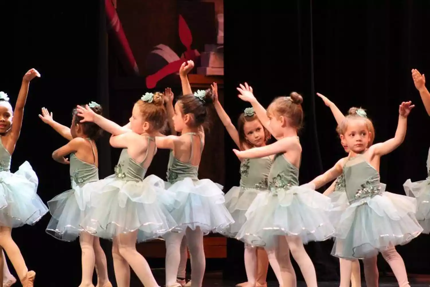 Young ballerinas in light green tutus perform on stage, with a backdrop featuring elements of The Nutcracker.