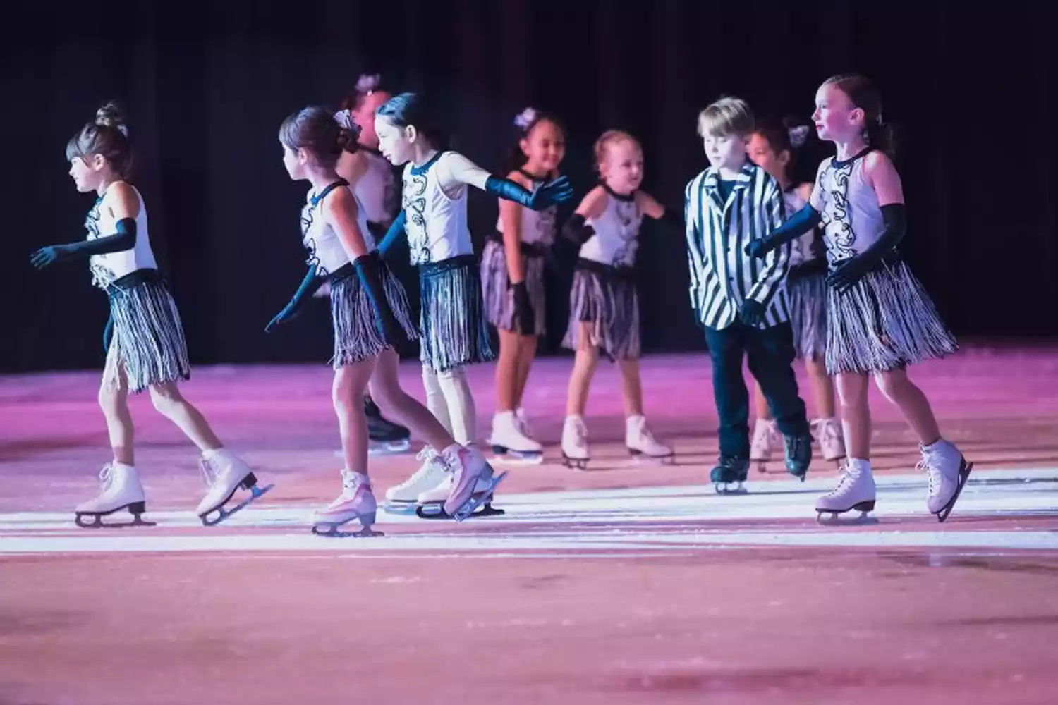 Children in matching costumes perform a synchronized ice skating routine on a brightly lit rink.