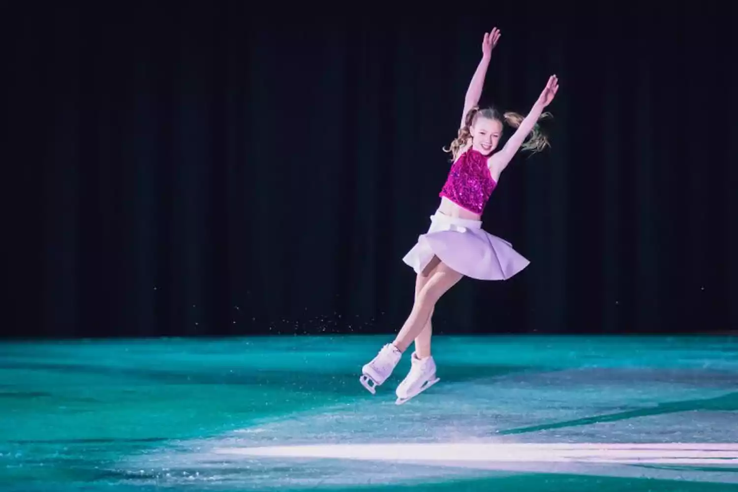 Young figure skater in a sparkly pink top and light purple skirt performs a jump on an ice rink, arms raised.