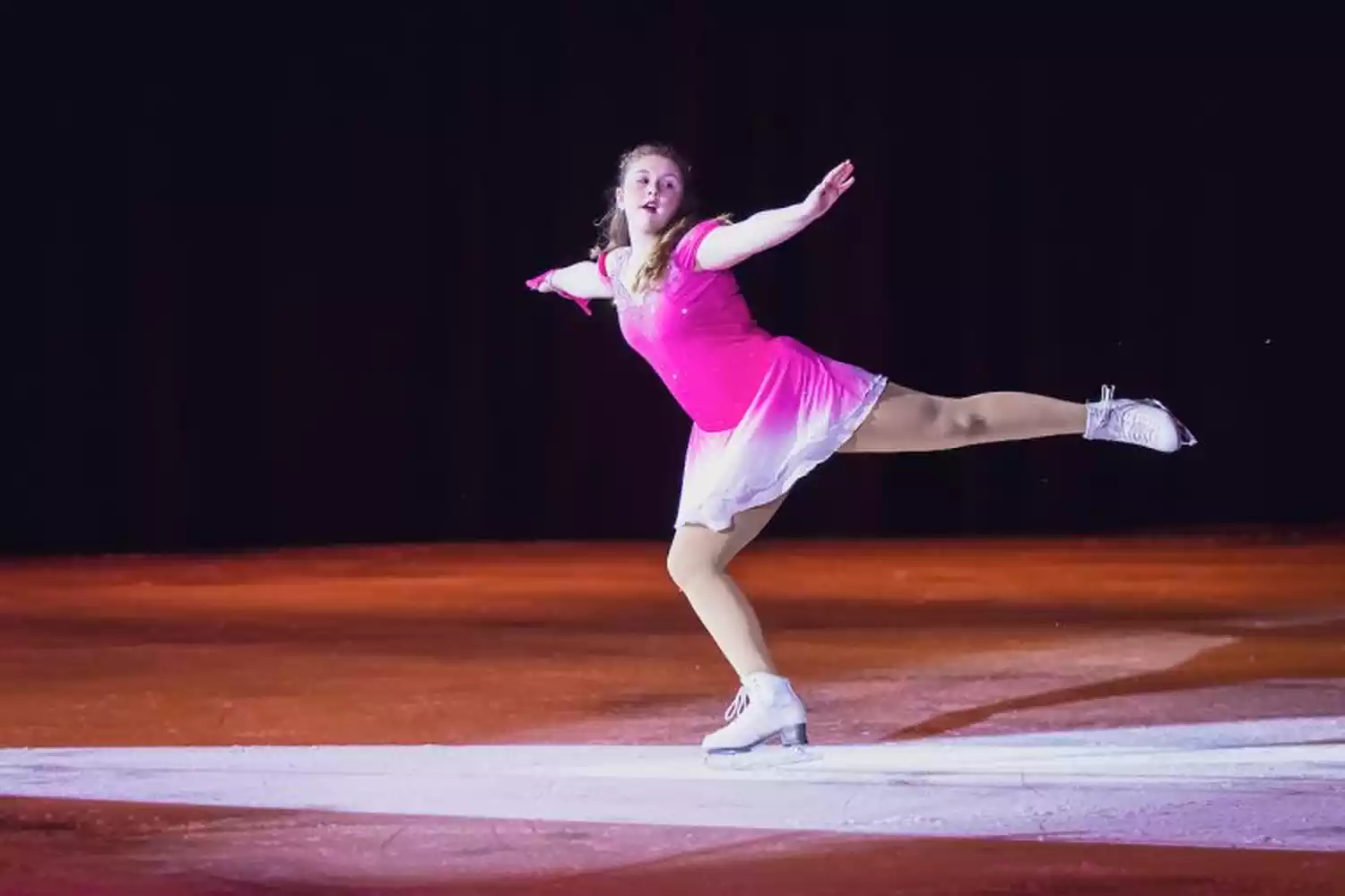 A female figure skater in a pink and white ombre dress performs a move on the ice during the Ice Show.