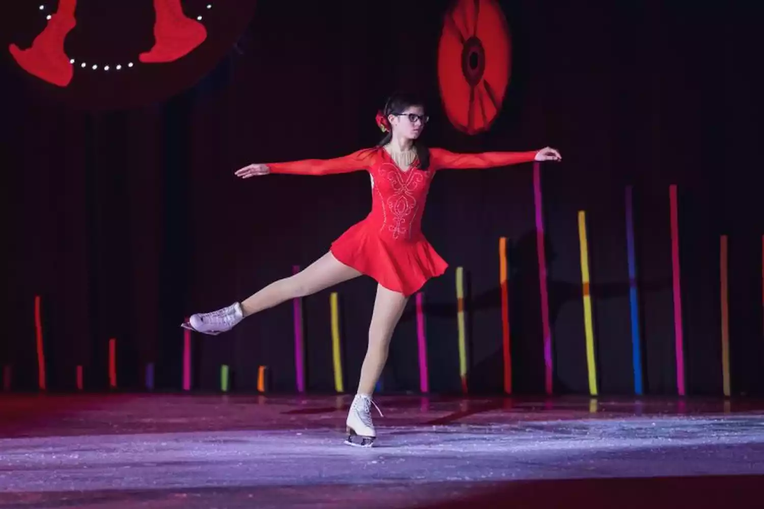 A figure skater in a red dress performs on ice, with arms outstretched and one leg extended. The background features colorful vertical lights and a