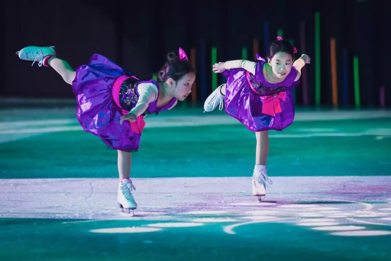 Two young girls in purple ice skating dresses perform on an ice rink, with colorful lights in the background.